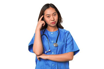 A young nurse chinese woman pointing temple with finger, thinking, focused on a task.