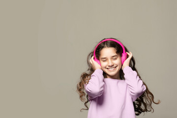 Energy girl with pink headphones listening to music with closed eyes on grey background in studio. She wears purple T-shirt, jeans. Long curly hair in tail is flying from moving.