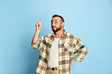 Portrait of bearded man in casual clothes, checkered shirt raising finger up, expressing ideas against blue studio background. Concept of human emotions, lifestyle, facial expression. Ad