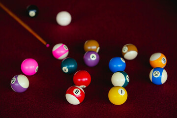 Assorted pool balls in various colors on a red fabric table, with a wooden pool cue next to it, illuminated with diffused light