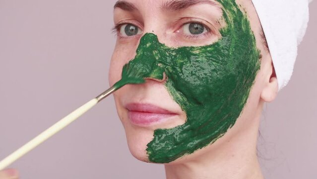 A young woman applies a green mask to her face with a brush. Facial skin care at home. Homemade face mask.	