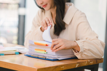 The busy work desk of a businesswoman is cluttered with stacks of paper files, folders, and papers as she searches and checks for unfinished documents, her hands moving swiftly over the paperwork.