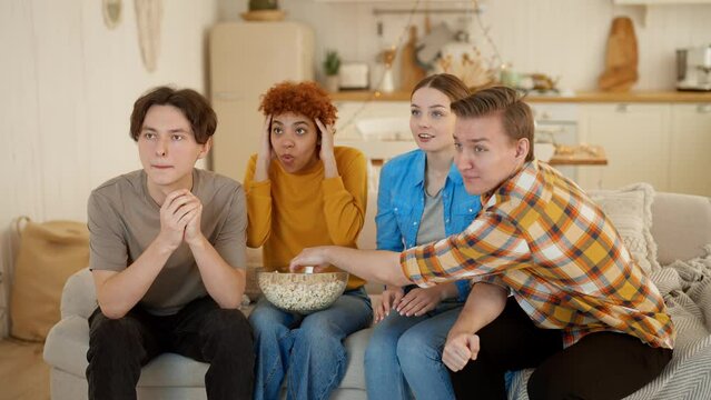 Company of diverse friends watches TV sports game and cheering favourite team. They emotionally talk, gesturing, waiting victory, rooting for team sitting on sofa at home eating popcorn together.