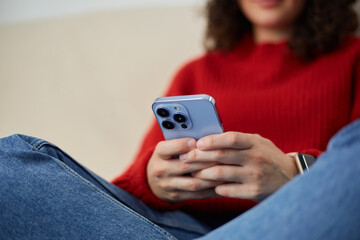 Young white woman typing a message on blue smartphone. Unrecognizable female person sitting on a couch at home and texting online with a mobile phone