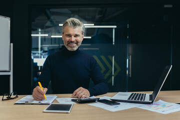 Portrait of a confident senior man businessman, accountant, manager sitting in the office at the table, smiling and looking at the camera. Work by laptop and with documents.