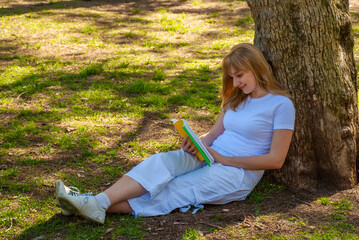 A girl with a book sits near a tree in the park