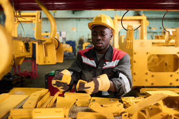 Young African American male engineer of factory choosing new spare parts for repairing industrial machine while standing against some of them