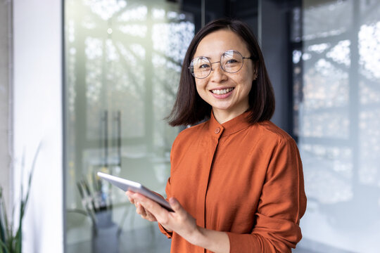 Portrait of a young successful Asian female programmer inside office, worker in glasses smiling and looking at the camera, a businesswoman holds a tablet computer in hands and uses it to test online .