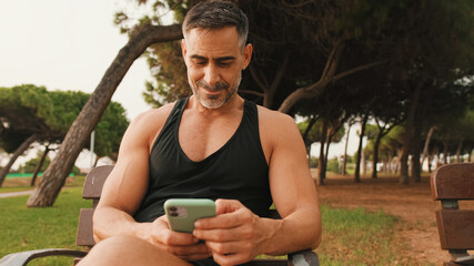 Smiling middle aged muscular man in sportswear using mobile phone while sitting in park after workout