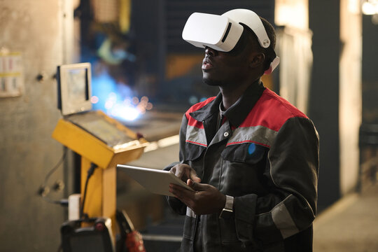 Young African American worker of factory in vr headset using tablet while operating industrial machine during technical production process