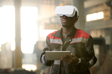 Young machine operator in vr headset and workwear using tablet while standing in front of machine and taking control of its work
