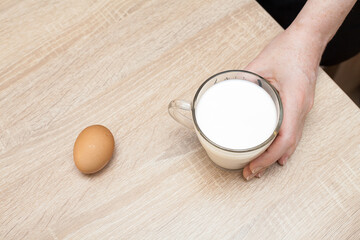 A woman's hand holds a glass of fresh milk on a textured table, a brown egg lies nearby. An image for your healthy food creatives.