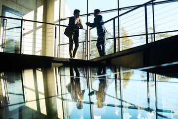 Outlines of two young male colleagues in formalwear standing by railing against large window in office center and discussing working points
