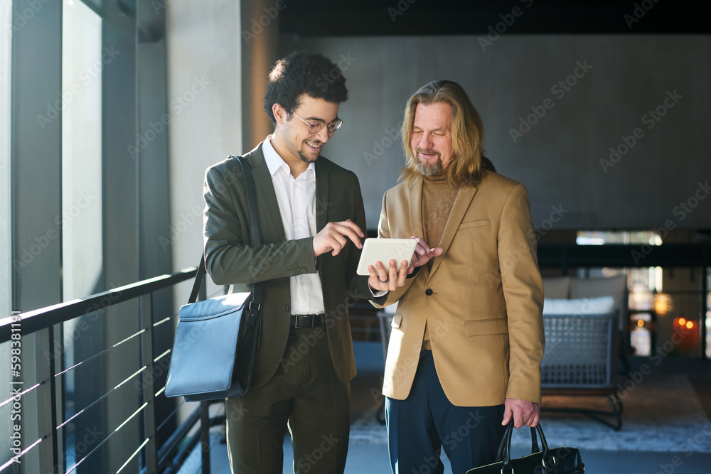 Aufkleber Young smiling broker with tablet scrolling through online data during presentation of updated information to mature male colleague