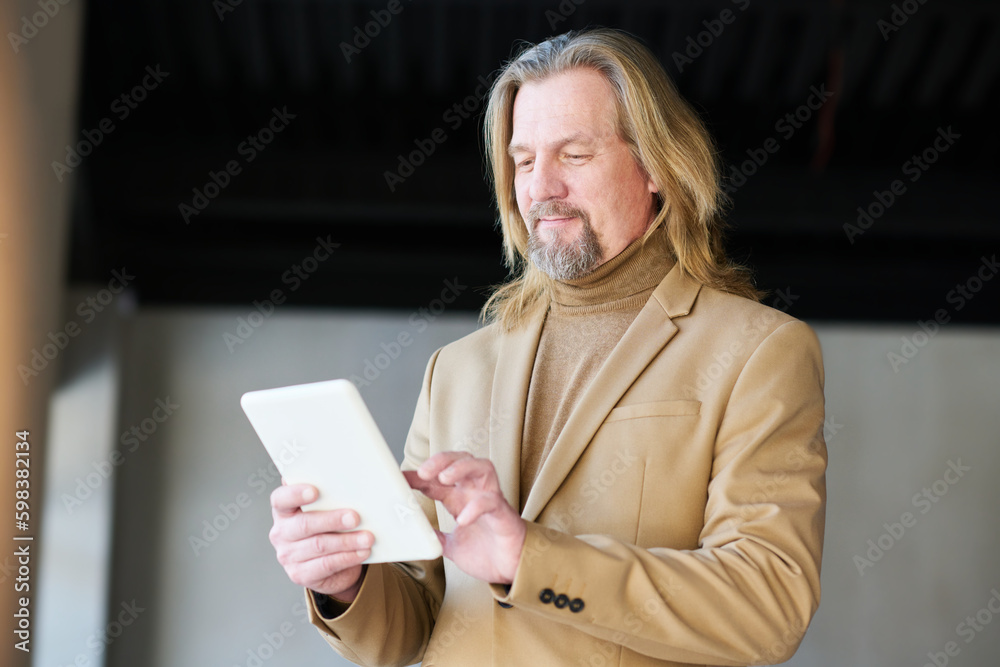 Poster Elegant mature businessman in beige formalwear using tablet while analyzing online graphic data or entering points of new project