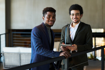 Two happy young intercultural male brokers in formalwear looking at camera while one of them using tablet to work with online data