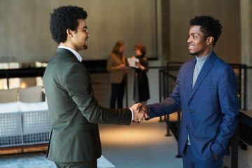 Side view of two young male business partners shaking hands at meeting or while greeting one another after negotiating and signing contract