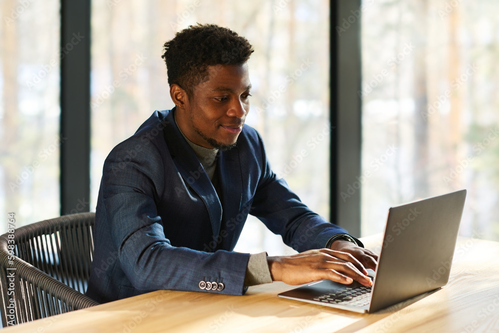 Aufkleber young smiling african american entrepreneur typing on laptop keyboard while sitting by workplace in 