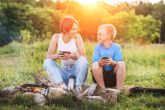 Smiling sister and brother kids have merry conversation near campfire. They looking eye to eye to each other, drinking tea from thermos. Happy family outdoor picnic camping activities concept