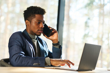 Young confident male broker or agent talking to client on smartphone while sitting by workplace in front of laptop and analyzing data