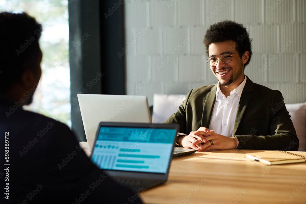 Poster happy young multi-ethnic businessman looking at colleague with laptop sitting in front of him during