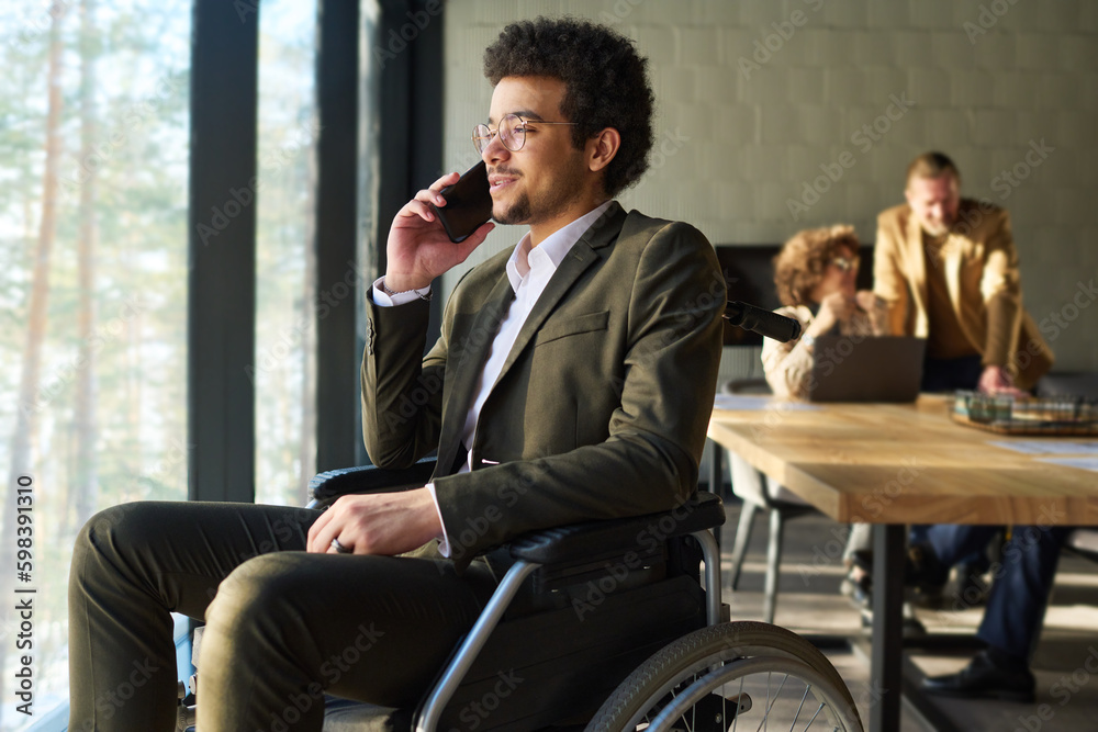 Aufkleber young confident businessman in whellchair sitting in front of window and consulting client on smartp