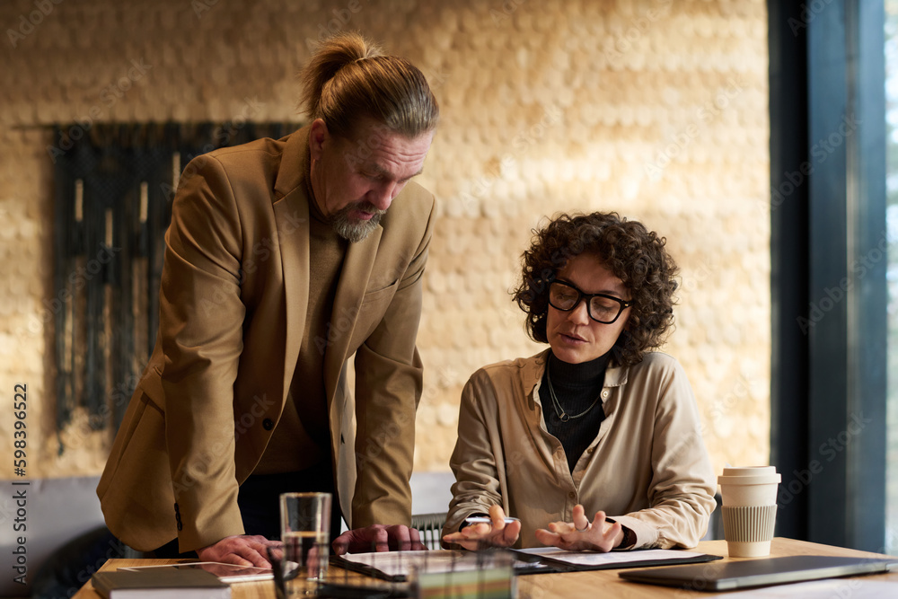 Poster confident businesswoman explaining data in document to mature male colleague standing by her and ben