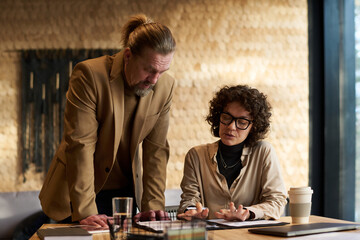 Fototapeta premium Confident businesswoman explaining data in document to mature male colleague standing by her and bending over table during discussion