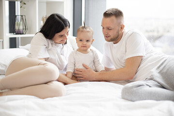 Obraz premium Caucasian parents lying on bed with baby and tenderly embracing from both sides. Sweet little girl with funny ponytail smiling lightly and looking ahead. Concept of love and family treasures.