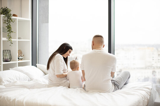 Back view of young parents holding cute infant while sitting on soft bed and enjoying panoramic view out of picture window. Cheerful adults and child relishing time together in bright apartment.