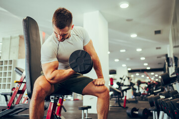 A focused sportsman is sitting in a gym and doing exercises for the biceps and triceps with a...