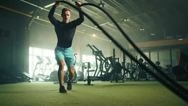 Process of doing split snatch waves on battle rope by young handsome man with fit body. Battle ropes workout. Fully equipped gym in the background. High quality 4k footage Generative AI
