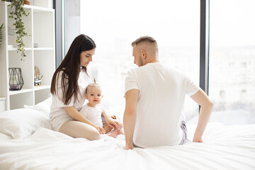 Obraz na płótnie Canvas Cute little baby girl sitting together with loving parents on soft duvet in bedroom during weekend at home. Cheerful caucasian mom and dad taking care of pretty daughter while relaxing indoors.