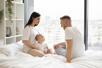 Fototapeta premium Cute little baby girl sitting together with loving parents on soft duvet in bedroom during weekend at home. Cheerful caucasian mom and dad taking care of pretty daughter while relaxing indoors.