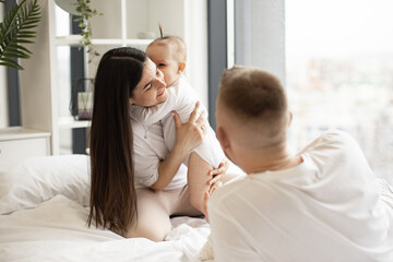 Obraz na płótnie Canvas Adorable baby infant hugging gently joyful brunette woman while happy blonde man lying on soft bed in studio apartment. Affectionate young family sharing love and closeness while living full life.