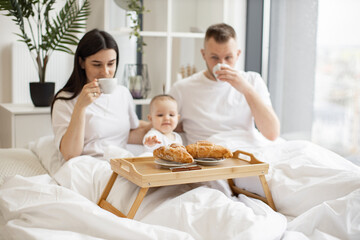 Obraz na płótnie Canvas Happy husband and wife holding coffee cups while cute kid reaching out for delicious food on bed tray in modern studio flat. Small caucasian family having enjoyable meal together in home interior.