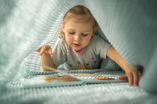 Little girl enjoying of review a book under blue knitted plaid in sunny morning Generative AI