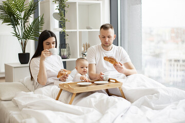 Obraz na płótnie Canvas Beautiful woman and handsome man sitting under duvet and having drinks and snacks while cute baby taking dessert from bed tray. Young parents and daughter enjoying excellent food in bright interior.