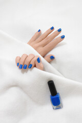 Woman hands with blue nails laing next to bottle of nail polish, on white blanket. Her hands are partially covered by white coverlet. Close up of blue manicure. Beauty concept.