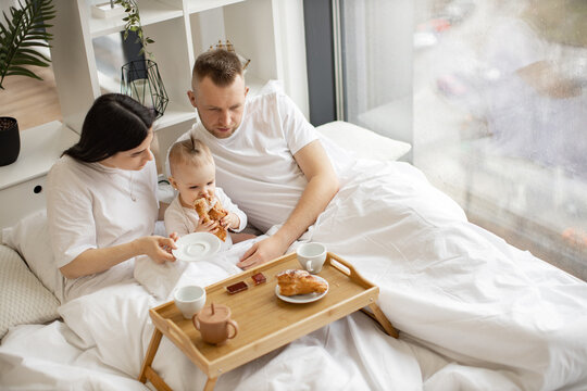 Top view of married caucasian couple helping baby girl eating roll while resting on bed with tray table in studio apartment. Loving young people taking care of cute daughter during morning time.