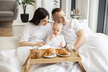 Sweet little baby drinking from sippy cup while joyful brunette woman snuggling with blonde male in cozy bedroom with tray on foreground. Caring family spending quality time together at home.