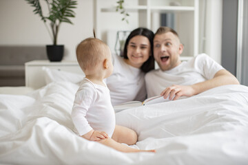 Fototapeta premium Focus on adorable little child sitting on white soft duvet and watching her parents playing roles of book characters on Sunday morning at home. Concept of family and togetherness.