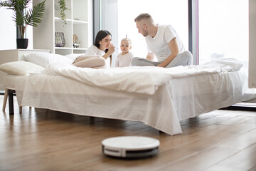 Relaxed spouses feeding small kid with spoon on bed while smart robotic vacuum removing dust from laminate floor in studio apartment. Modern parents with baby using electronic housekeeping technology.