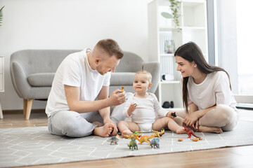 Fototapeta na wymiar Young married couple sitting cross-legged in living room and playing dinosaur game with little infant daughter on carpet. Loving family of three spending quality time together at home.