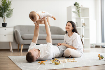 Caucasian father lifting baby girl up high in air while lying among toys on floor near smiling...
