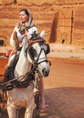 Naklejka na ściany i meble Young woman with white dress and head scarf, sitting on a horse, blurred Hegra or Mada’in Salih ruins in background - female tourist travel influencer like photo from Saudi Arabia