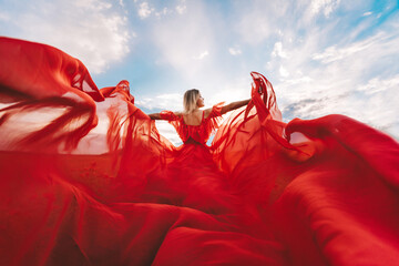 Woman Red Flying Dress. A blonde in a red dress against the sky. Rear view of a beautiful blonde woman in a red dress fluttering in the wind against a blue sky and clouds. - obrazy, fototapety, plakaty