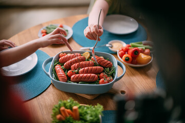 A dish with baked sausages and vegetables on the table and children's hands with forks. Healthy eating for children.