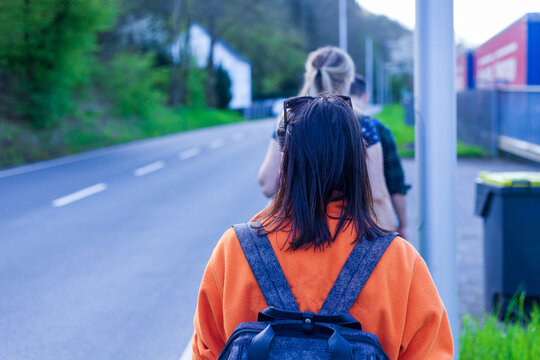 Back View Of Young Woman With Backpack Walking On The Street. Rear View Of Young Woman In Orange Jacket And Black Sunglasses Walking On The Street