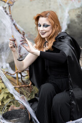 Naklejka premium Attractive beautiful woman in a Halloween costume black cloak and scary make-up. Young girl in a look of a witch dressed up for a party making fairy potion holding black lantern